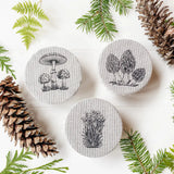three mini mason jar covers with a mushroom design surrounded by pine branches and ferns