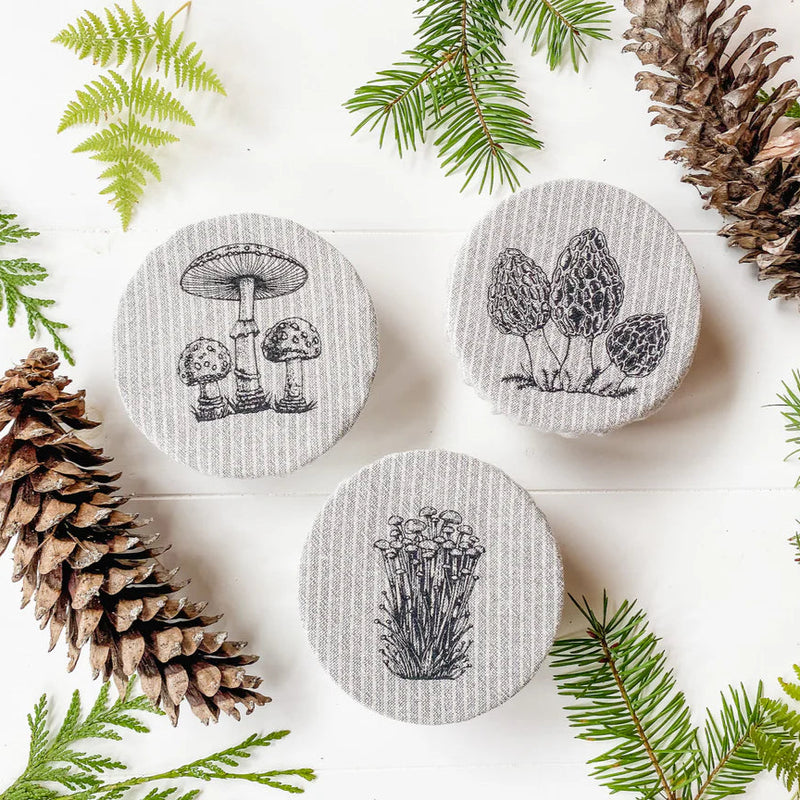 three mini mason jar covers with a mushroom design surrounded by pine branches and ferns